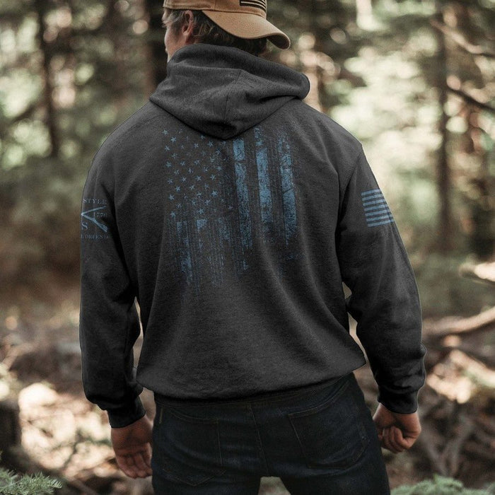 Person wearing a dark hoodie with a flag design in a forest setting