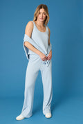 A person stands against a blue background wearing the P.J. Salvage 3-Piece Luxe Ribbed Celestial PJ Set: a sleeveless light blue ribbed top, matching wide-leg pants, and a long-sleeve top draped over their elbows with white shoes.
