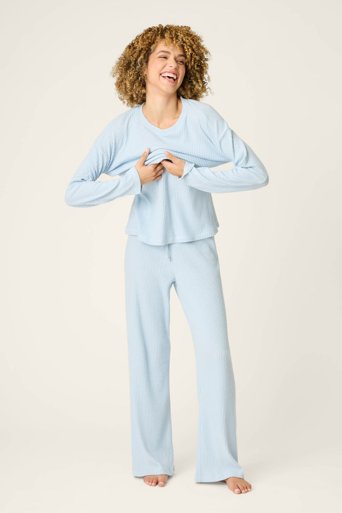A person with curly hair models the P.J. Salvage 3-Piece Luxe Ribbed Celestial PJ Set in light blue, standing barefoot on a neutral background, smiling and holding the hem of their shirt.