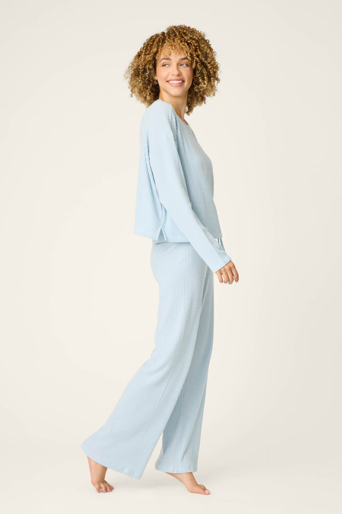 A person with curly hair is wearing the P.J. Salvage 3-Piece Luxe Ribbed Celestial PJ Set in light blue, featuring a long-sleeve top and wide-leg pants, standing barefoot and smiling against a plain light background.