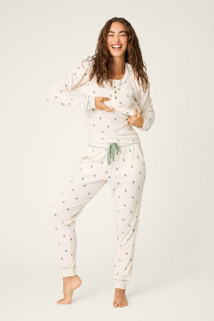 A person with long brown hair smiles in the P.J. Salvage 4-Piece Tree Print Thermal Lounge Set, standing barefoot on one leg and holding their shirt’s hem, radiating cozy cabin getaway vibes.