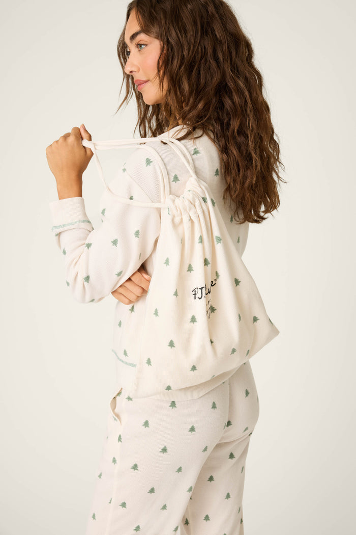 A woman with wavy brown hair wears the P.J. Salvage 4-Piece Tree Print Thermal Lounge Set, a cream winter pajama set with a green tree pattern, and carries a matching drawstring bag—perfect for a cozy cabin getaway.