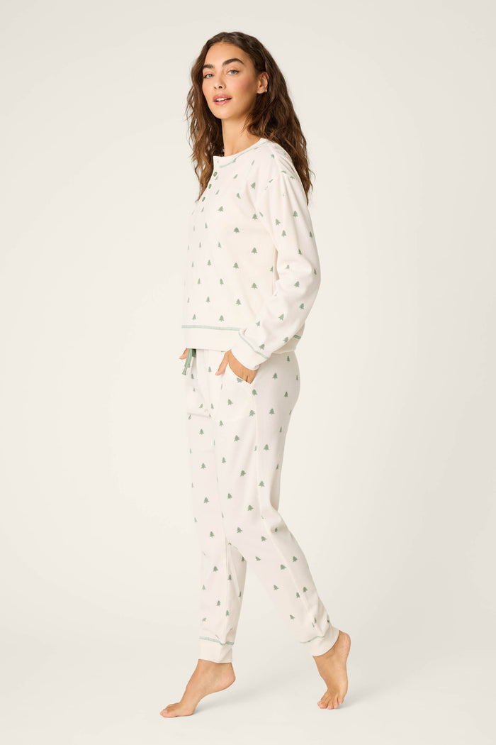 A person stands barefoot against a plain light background, modeling the P.J. Salvage 4-Piece Tree Print Thermal Lounge Set—a matching white pajama set with small green tree prints, including long sleeves and jogger-style pants.