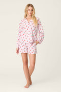 A woman stands barefoot on a plain background in the Afterparty Poplin Oh Cherry Short PJ Set by P.J. Salvage—a powder pink cotton poplin pajama set with a colorful cherry print, long-sleeve button-up top, and matching shorts. One hand rests in her pocket.