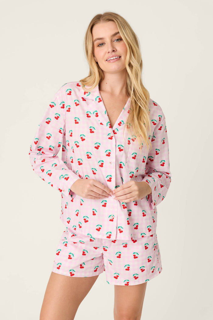 A woman with long blonde hair models the P.J. Salvage Afterparty Poplin Oh Cherry Short PJ Set, featuring a cotton poplin long-sleeve button-up top and matching shorts in a gingham print with cherry motifs, against a light background.