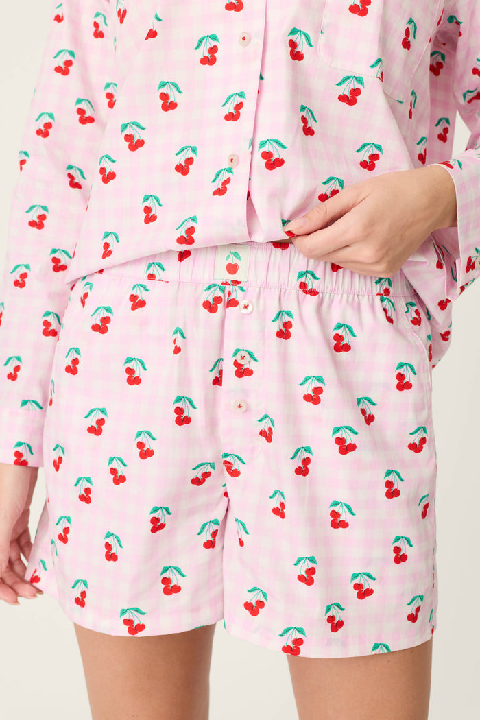 A person wears the Afterparty Poplin Oh Cherry Short PJ Set by P.J. Salvage in powder pink cherry print cotton poplin. The image highlights the waist and upper thigh, with a hand softly adjusting the shorts’ waistband.