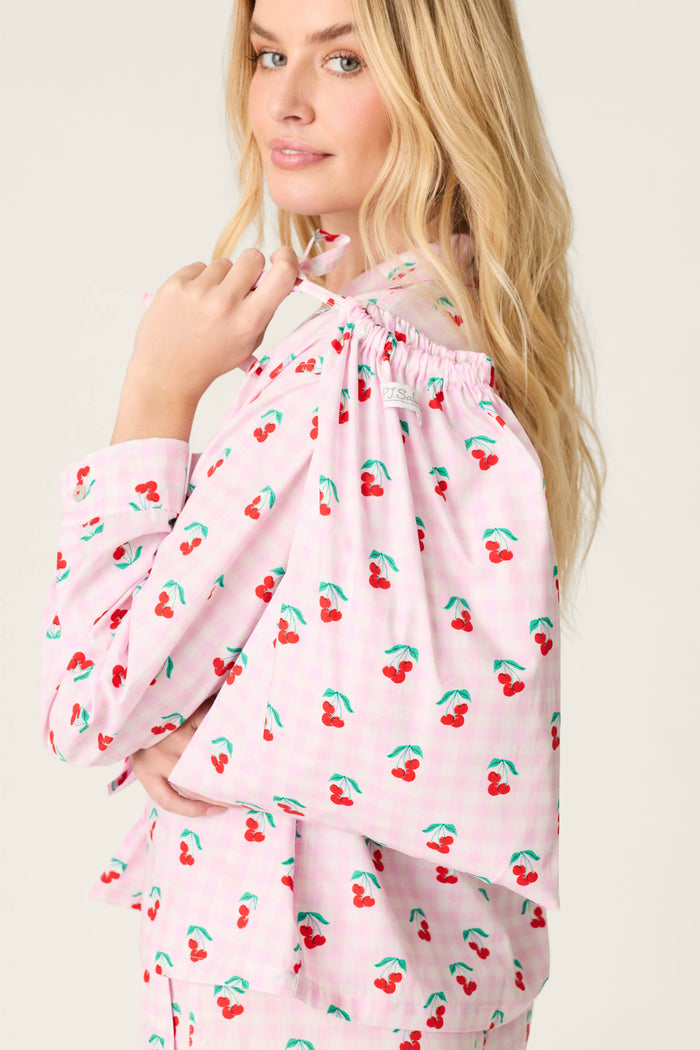 A woman with long blonde hair models the P.J. Salvage Afterparty Poplin Oh Cherry Short PJ Set, featuring a cherry print and matching cape, as she looks over her shoulder against a plain, light background.