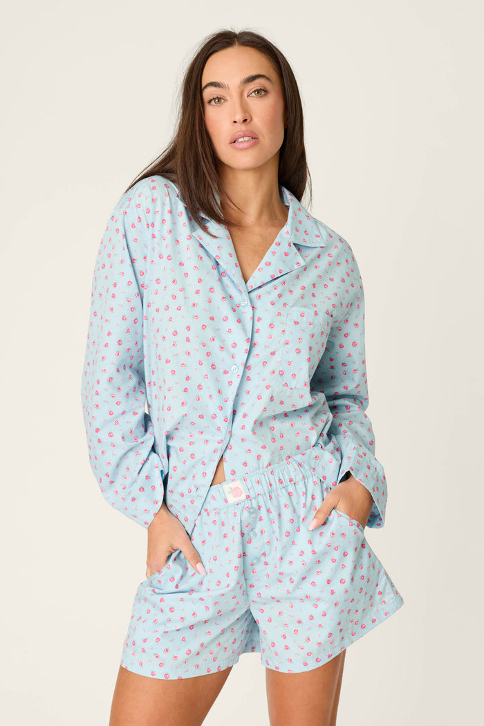 A person with long brown hair stands against a plain light background wearing the Afterparty Poplin Rosy Nights Short PJ Set by P.J. Salvage—a light blue cotton poplin set with a delicate rose print, featuring a button-up top and matching shorts.