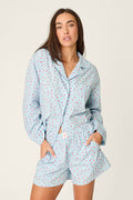A person with long brown hair stands against a plain light background wearing the Afterparty Poplin Rosy Nights Short PJ Set by P.J. Salvage—a light blue cotton poplin set with a delicate rose print, featuring a button-up top and matching shorts.
