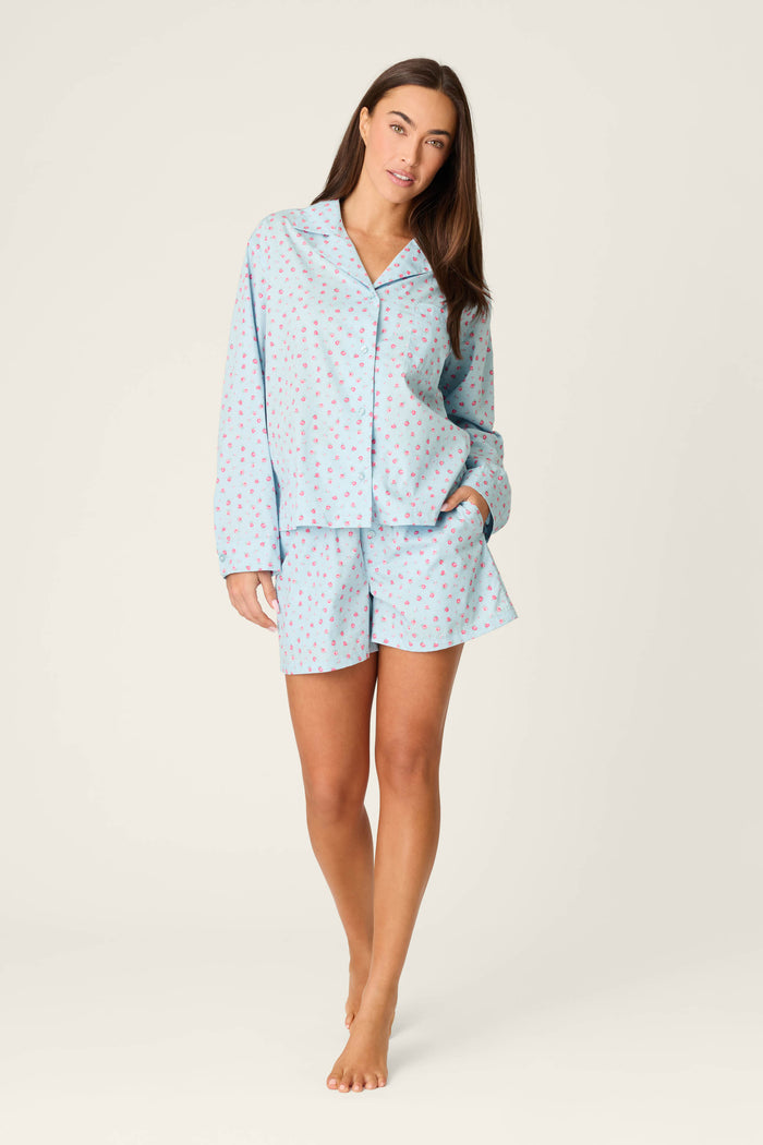 A woman with long brown hair stands barefoot on a plain background wearing the P.J. Salvage Afterparty Poplin Rosy Nights Short PJ Set—a light blue cotton poplin pajama set with long sleeves, shorts, and a pink and blue spot pattern. One hand is in her pocket.