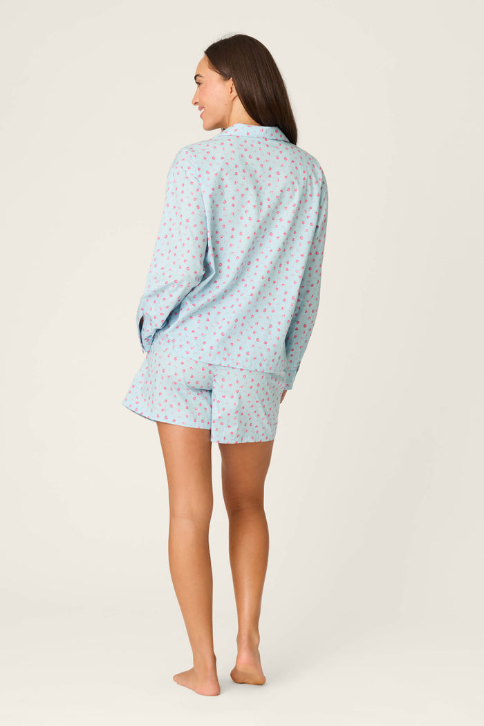 A person stands barefoot, facing away, wearing the P.J. Salvage Afterparty Poplin Rosy Nights Short PJ Set—a blue cotton poplin pajama set with a collared long-sleeve top and patterned shorts—against a plain light background.