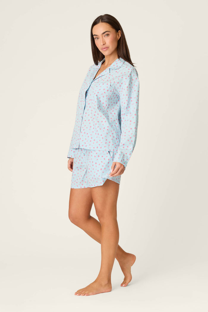 A woman stands barefoot in the Afterparty Poplin Rosy Nights Short PJ Set by P.J. Salvage, featuring a soft cotton poplin long-sleeve button-up top and matching shorts with a red and blue heart pattern, against a plain light background.