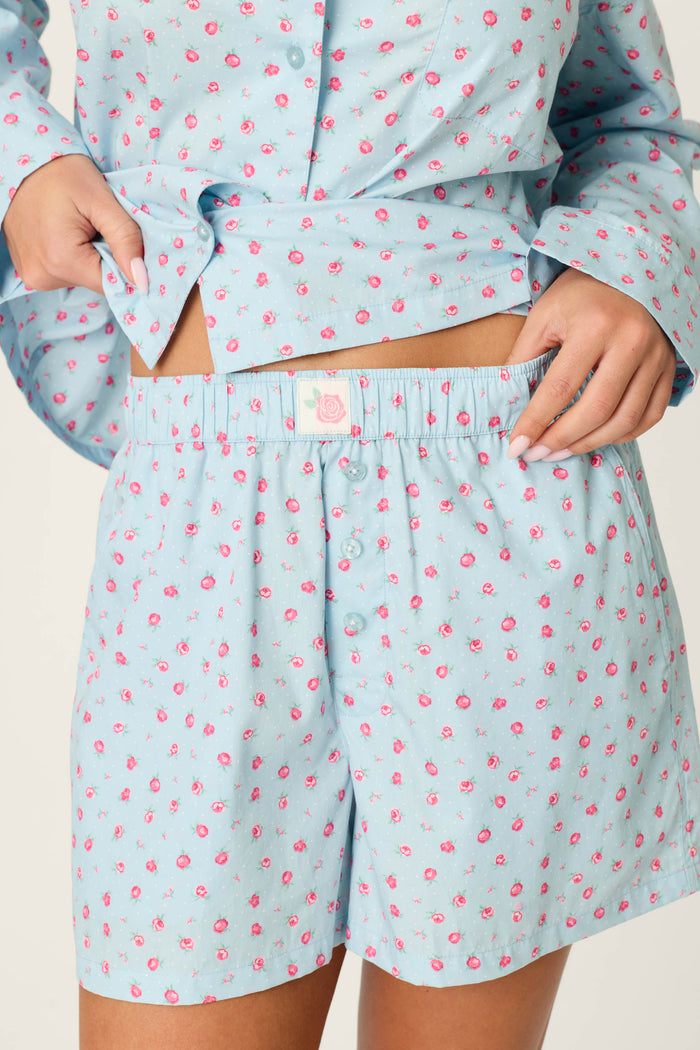 A person adjusts the waistband of the Afterparty Poplin Rosy Nights Short PJ Set by P.J. Salvage, featuring blue cotton poplin with a small pink rose print. The image focuses on their midsection and hands.