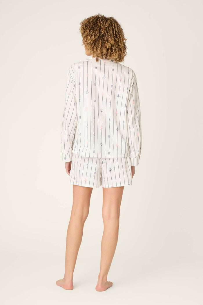 A person stands barefoot facing away from the camera, wearing the P.J. Salvage Afterparty Poplin Anchors Away Short PJ Set—an ivory cotton poplin pajama set with a long-sleeve shirt and matching vertically striped, dotted shorts—against a plain, light background.