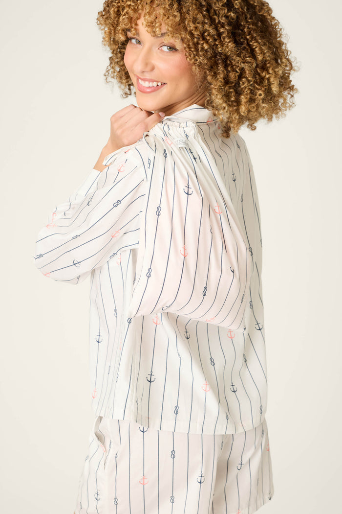 A woman with curly hair smiles over her shoulder, wearing the P.J. Salvage Afterparty Poplin Anchors Away Short PJ Set—an ivory, long-sleeved top and matching striped anchor-print shorts. The look is coastal and relaxed against a plain light background.