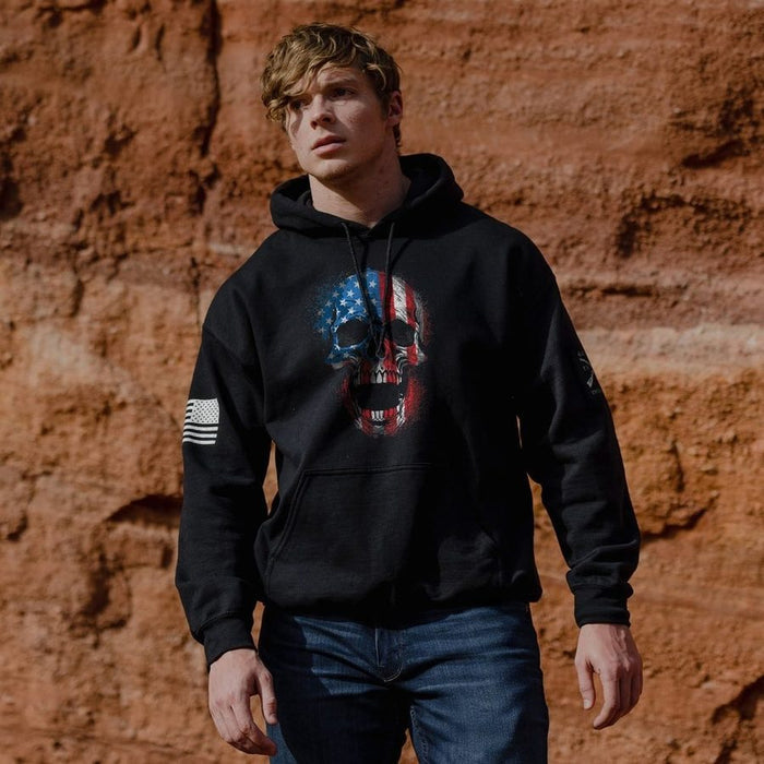 Person wearing a black hoodie with a skull design and American flag, standing against a red-brown brick wall.