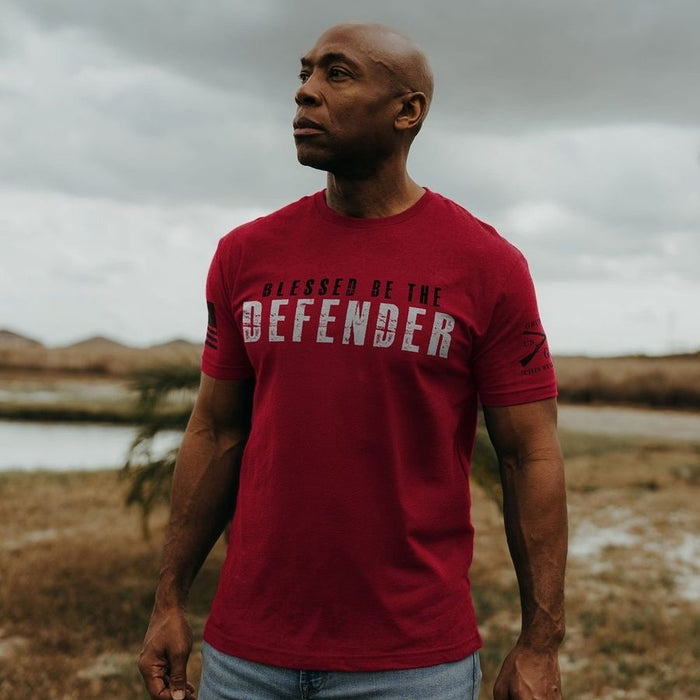 Man wearing a red t-shirt with 'Blessed be the Defender' text outdoors.