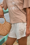 A person in a beige AMORE seashell tee, P.J. Salvage Breeze By The Sea White Sands Shorts, and holding a woven straw handbag stands by a green railing near water.