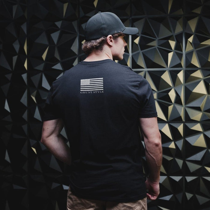 Man wearing a black t-shirt with an American flag design on the back, standing against a geometric patterned wall.