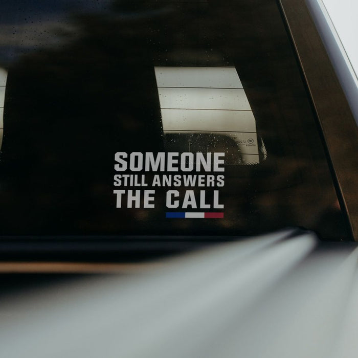 A white vinyl decal with the text 'SOMEONE STILL ANSWERS THE CALL' and a small blue and red accent bar, applied to the rear window of a vehicle.