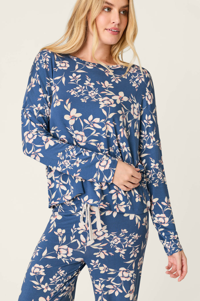 A woman wears the P.J. Salvage Flora Dreams Twilight Garden Long Sleeve PJ Top, featuring a blue background with a light floral print, paired with matching pants and standing against a plain light backdrop.