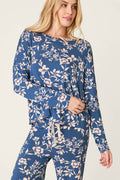 A woman wears the P.J. Salvage Flora Dreams Twilight Garden Long Sleeve PJ Top, featuring a blue background with a light floral print, paired with matching pants and standing against a plain light backdrop.