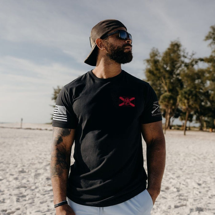 A man wearing a black t-shirt with a red chest logo and a black baseball cap, standing on a sandy beach with trees in the background.