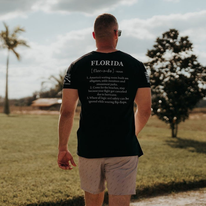 A man viewed from behind wearing a black t-shirt with white printed text on the back and light beige shorts, standing outdoors in a grassy field under a cloudy sky.