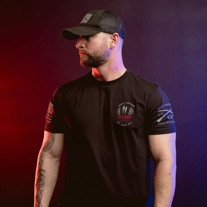A man wearing a black short-sleeve t-shirt with a circular '25 Years' logo on the left chest, a black trucker hat, and tattoos on his arm, posed against a dark background with red and blue lighting.