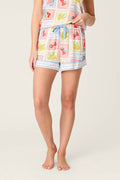 A person stands barefoot with their left hand in the pocket of P.J. Salvage Frutta Fresca Summer Harvest Camisole shorts, featuring a drawstring waist and red, yellow, green, and blue floral, leaf, and fruit prints. Upper body partly out of frame.