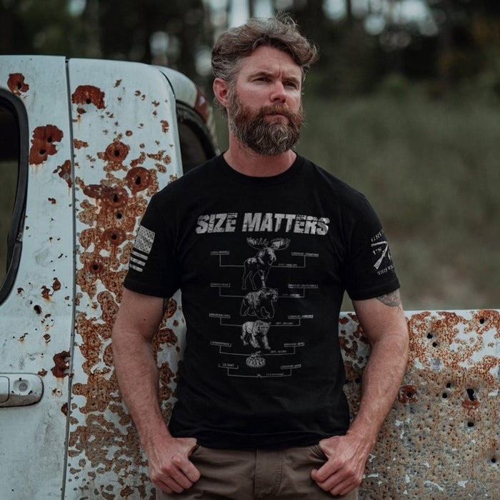 A bearded man wearing a black graphic t-shirt with a moose size chart and the words 'SIZE MATTERS' on the front, standing outdoors against a rusted white truck.