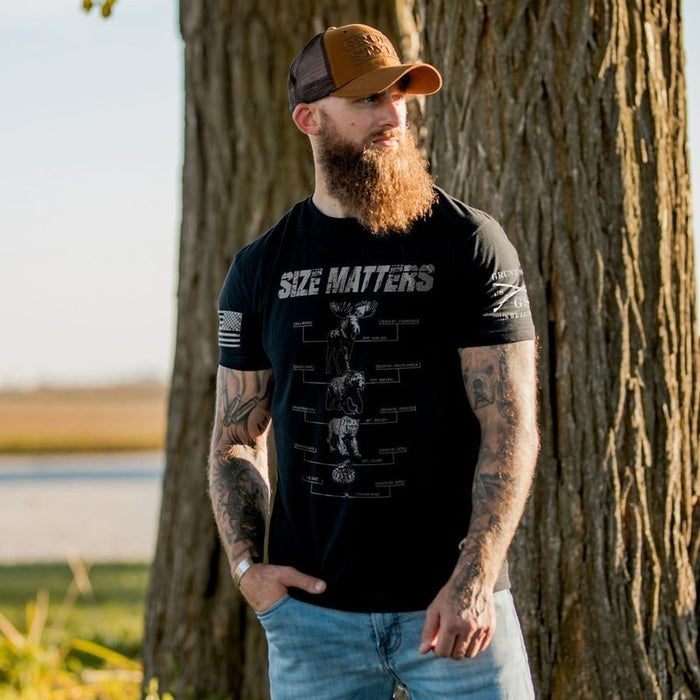 A bearded man wearing a black graphic t-shirt with 'SIZE MATTERS' printed on the front, paired with a brown and black trucker hat and light blue jeans, standing outdoors next to a large tree trunk.