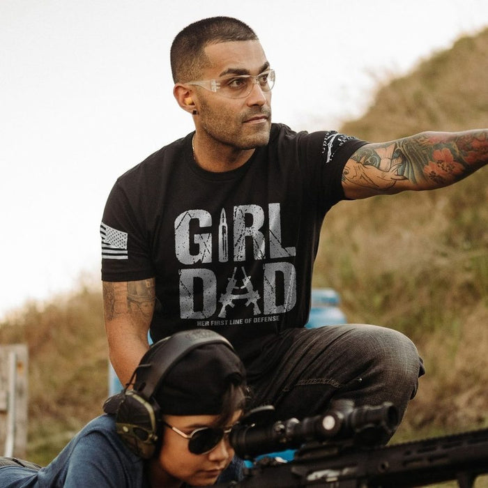 Man wearing a black t-shirt with 'GIRL DAD' text, sitting outdoors with a child and rifle.