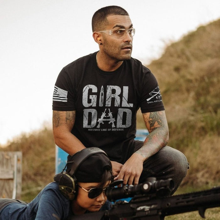 Man wearing a black t-shirt with 'Girl Dad' text, sitting outdoors with a child and rifle.