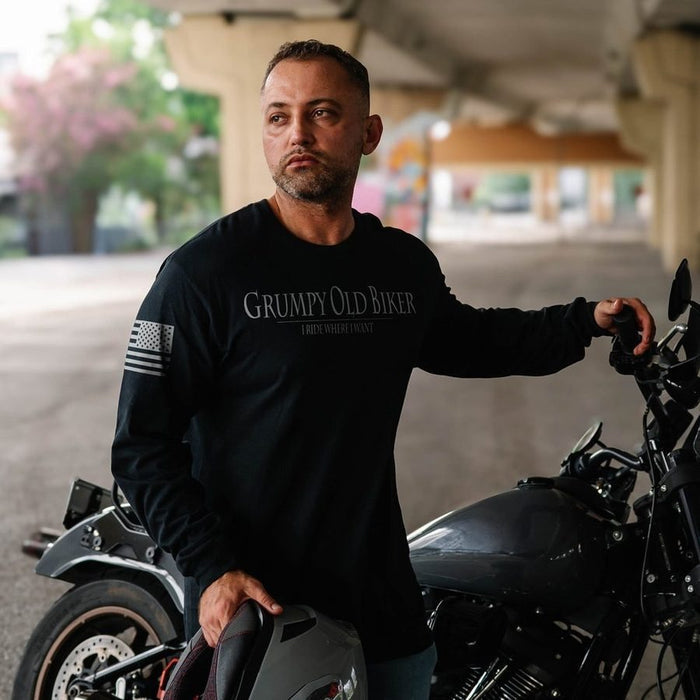 A man wearing a black long-sleeve t-shirt with 'Grumpy Old Biker' printed on the chest stands next to a motorcycle in an outdoor underpass setting.