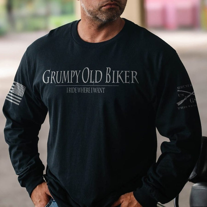 A man wearing a black long-sleeve t-shirt with 'GRUMPY OLD BIKER I RIDE WHERE I WANT' printed in white on the chest, featuring a distressed American flag on the right sleeve and a graphic on the left sleeve.