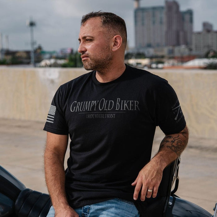A man wearing a black graphic t-shirt with 'Grumpy Old Biker' printed on the front, sitting outdoors next to a vehicle with a concrete wall and city buildings in the background.
