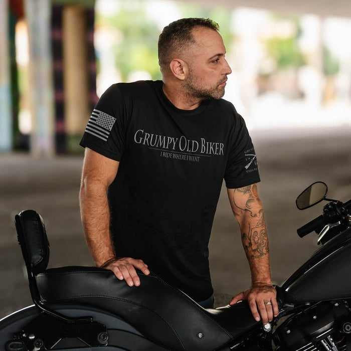 A man wearing a black graphic t-shirt with 'Grumpy Old Biker' printed on the chest, leaning on a black motorcycle in a blurred outdoor setting.