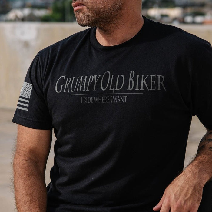 A man wearing a black short-sleeve t-shirt with 'Grumpy Old Biker' and 'I RIDE WHERE I WANT' printed in gray on the chest, plus a distressed American flag graphic on the sleeve.