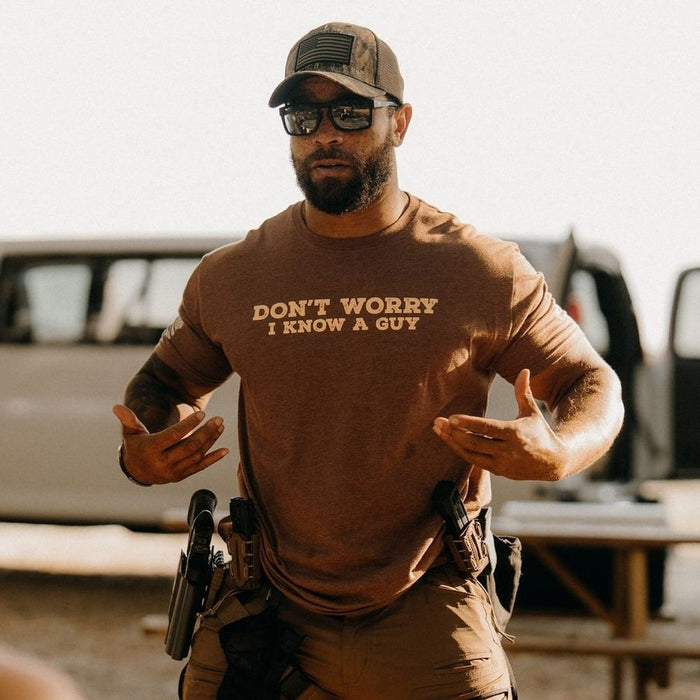 Man wearing a brown t-shirt with 'Don't Worry, I Know A Guy' text, standing outdoors.