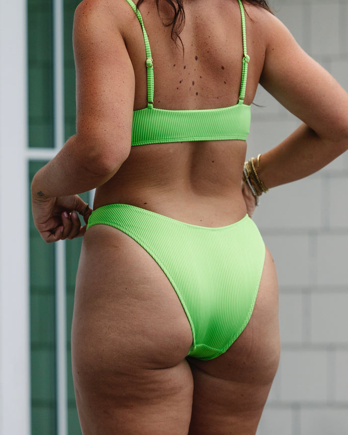 A woman wearing Honey Swim Kiwi Ribbed Bottoms stands outdoors with her back to the camera near a white building and glass window, showing her arms, back, and legs.