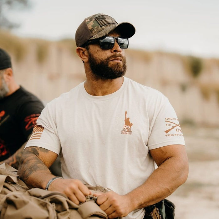 Man wearing a white t-shirt with logos, sunglasses, and a cap in an outdoor setting.