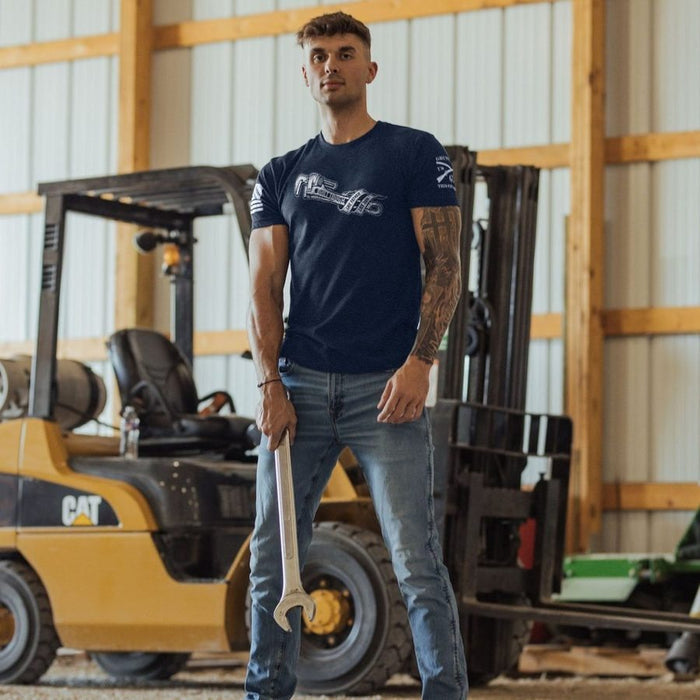 Man standing in a warehouse with forklifts, wearing a navy blue t-shirt and jeans.