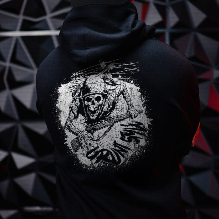 A person shown from the back wearing a black hoodie with a large distressed white graphic on the back featuring a skull, barbed wire, and a rifle. The background is a dark geometric wall with red accents.
