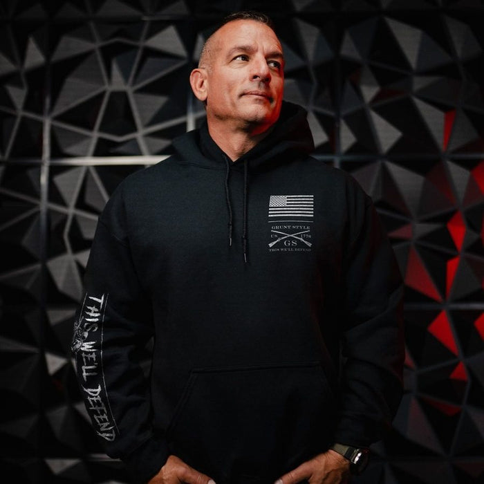 A man wearing a black pullover hoodie with a white American flag and 'Guns & Ammo' logo on the left chest, and 'THIS WILL DEFEAT' text on the right sleeve, posed against a dark geometric wall.