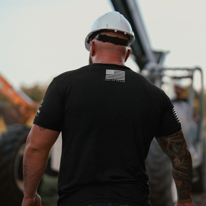 Man wearing a black t-shirt with an American flag design and a hard hat, standing in front of construction equipment.