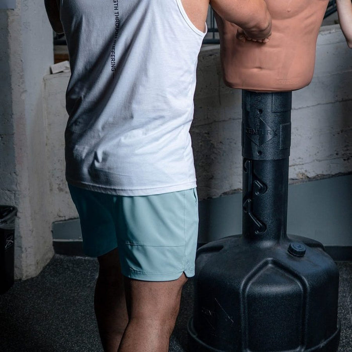 Person wearing a white tank top and light blue shorts standing next to a large black container in an indoor setting.