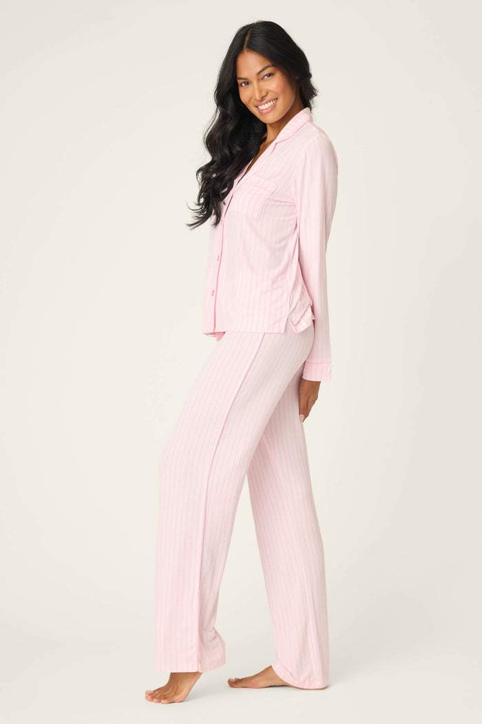 A person with long black hair smiles barefoot in the P.J. Salvage Modal Basics Pink Stripe 3-Piece PJ Set, featuring a light pink striped button-up top and loose pants, against a plain background.
