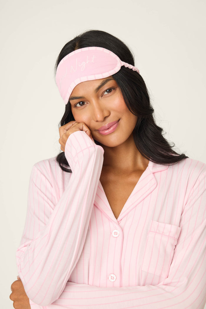 A woman smiles gently, wearing the P.J. Salvage Modal Basics Pink Stripe 3-Piece PJ Set and a matching pink sleep mask with cursive script, posing against a plain background.