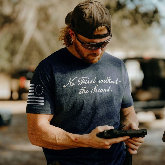 Man wearing a t-shirt with text and a flag design, holding a gun outdoors.